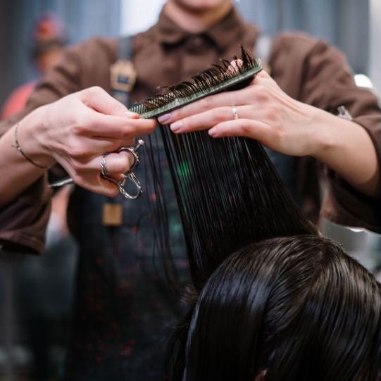 A hairstylist's hands comb a person's wet hair, preparing to cut it with a pair of scissors dangling from a finger on one of their hands.