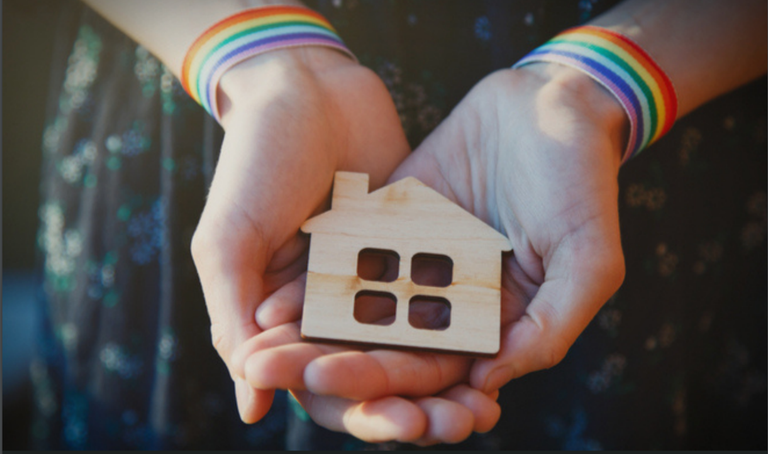 a pair of hands wearing rainbow bracelets hold a wooden cutout shaped like a side profile of a home with a square window.