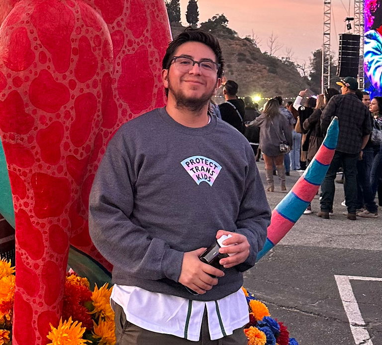A man of brown complexion with short, dark hair smiles kindly as he stands in front of a large, red sculpture at a crowded event. He is wearing a gray shirt that says Protect Trans Kids.