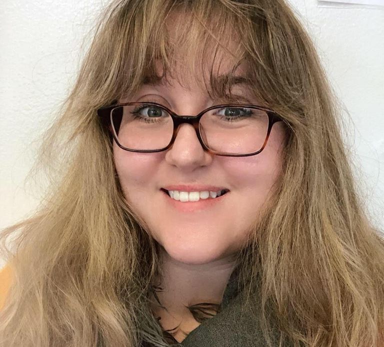 A young woman of fair complexion with long blonde hair and glasses smiles shyly at the viewer