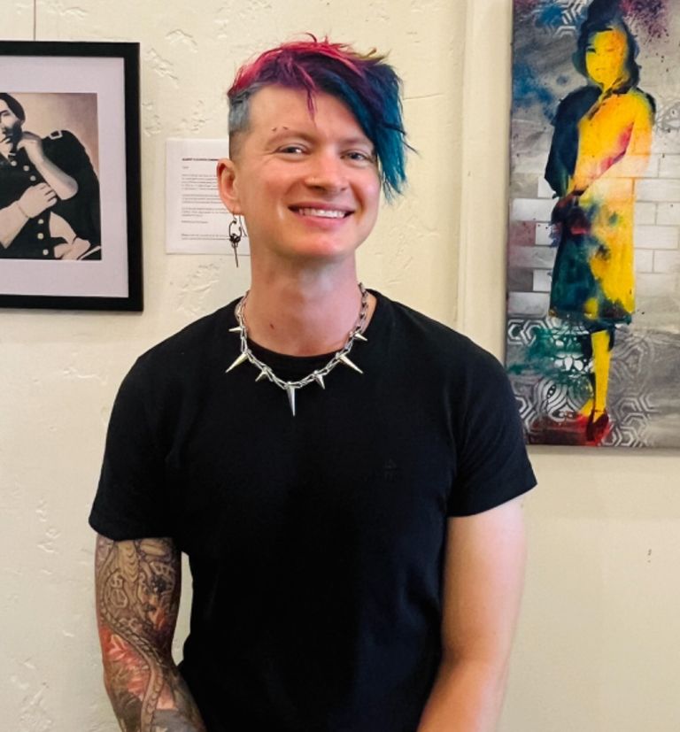 A slender man of fair complexion with short, rainbow hair and a full sleeve of tattoos wearing a black shirt and silver necklace and earrings smiles as he stands between a colorful spray-painted painting and a black and white vintage-styled painting.